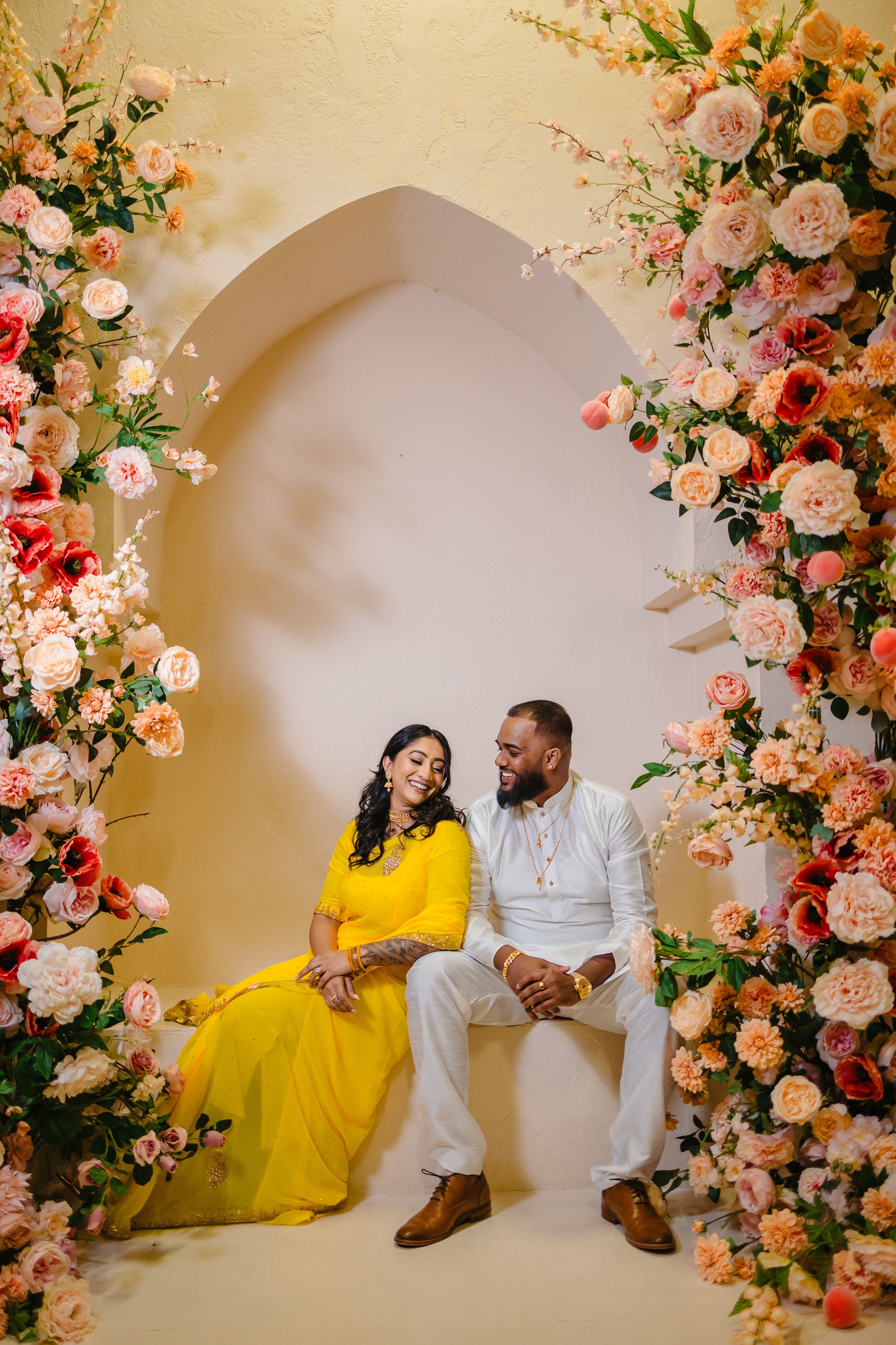 Couple posing under an elegant floral arch with yellow dress and white attire.