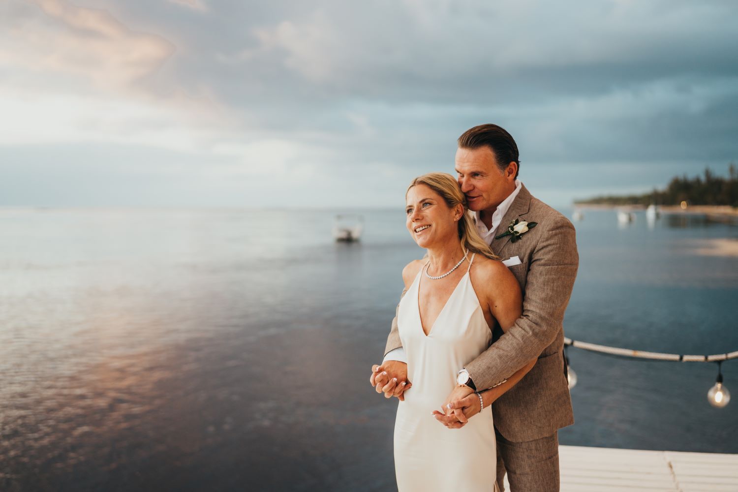 Brautpaar vor einem Sonnenuntergang mit dem Meer im Hintergrund. Die Braut trägt ein weisses Brautkleid mit V-Ausschnitt.