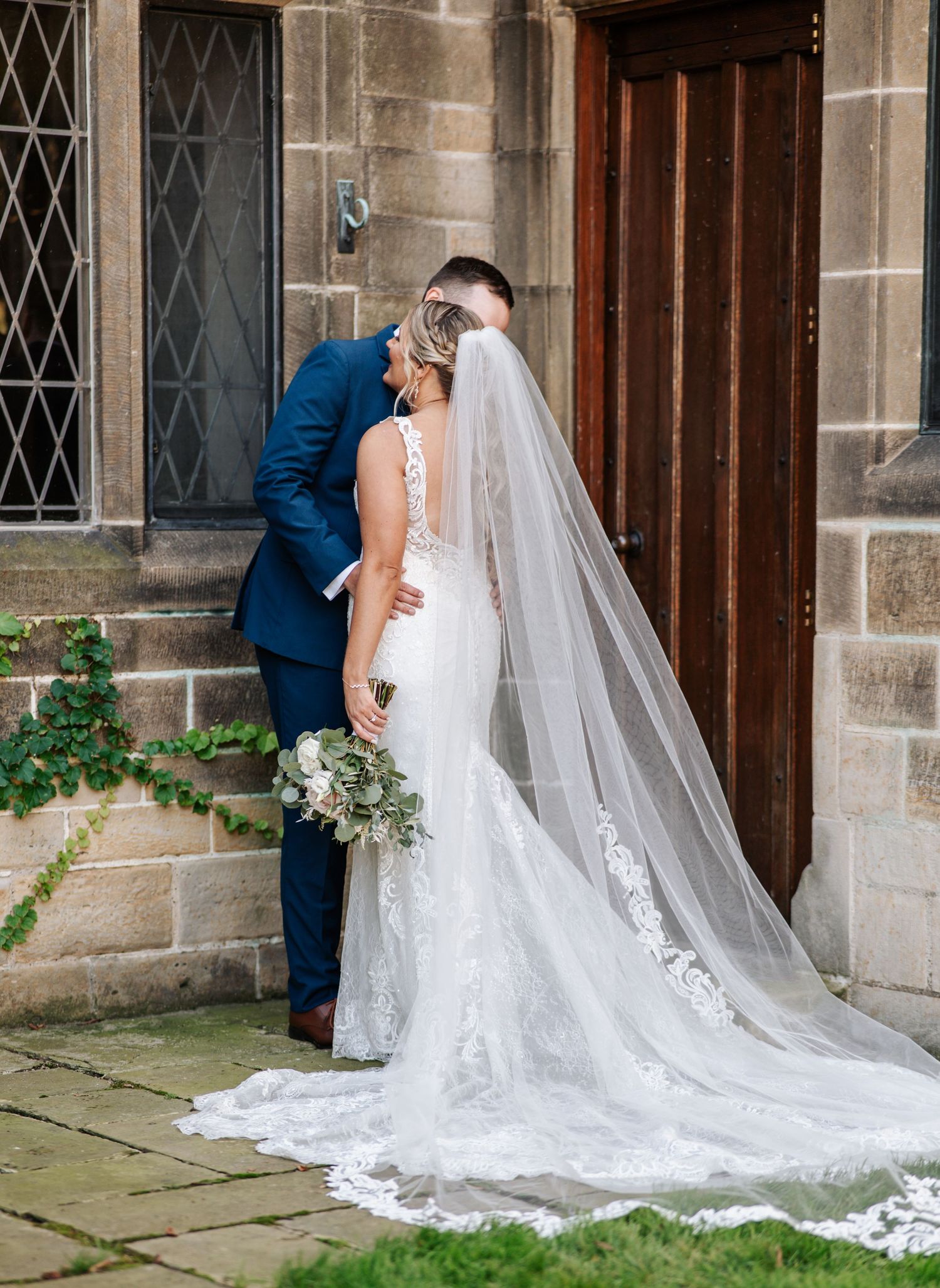 Elegant wedding couple poses outside historic church with flowing white lace veil trailing on stone steps.