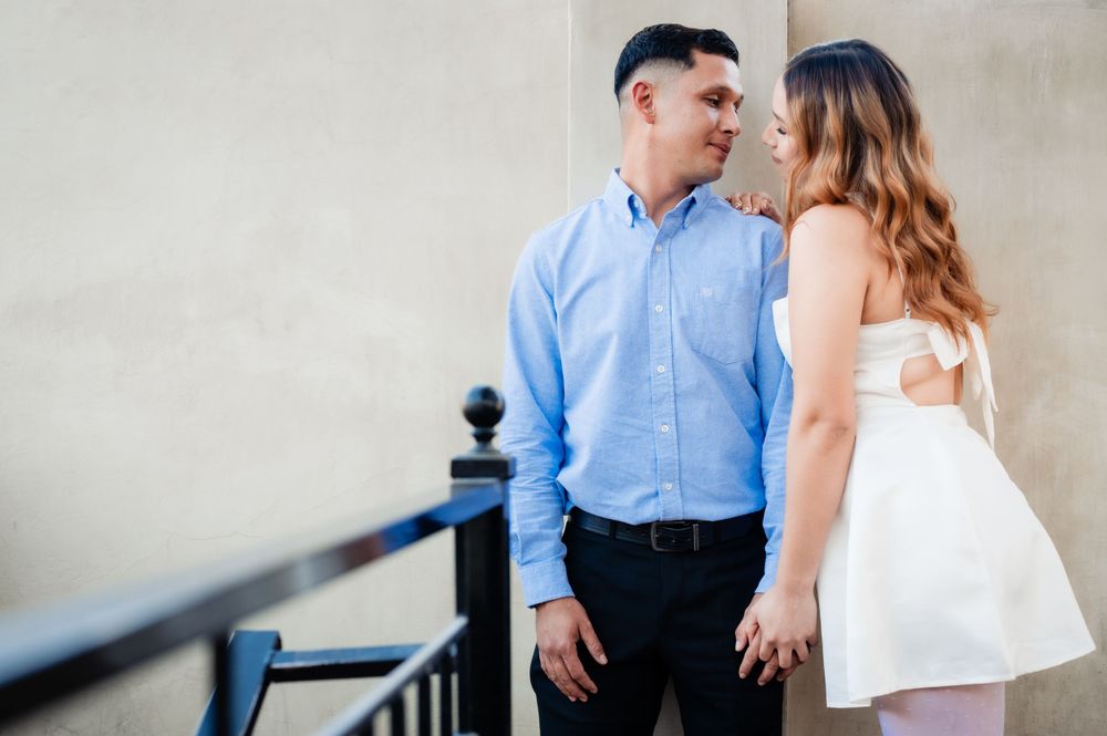 A couple in formal attire gazes at each other while standing against a neutral wall near a staircase railing.