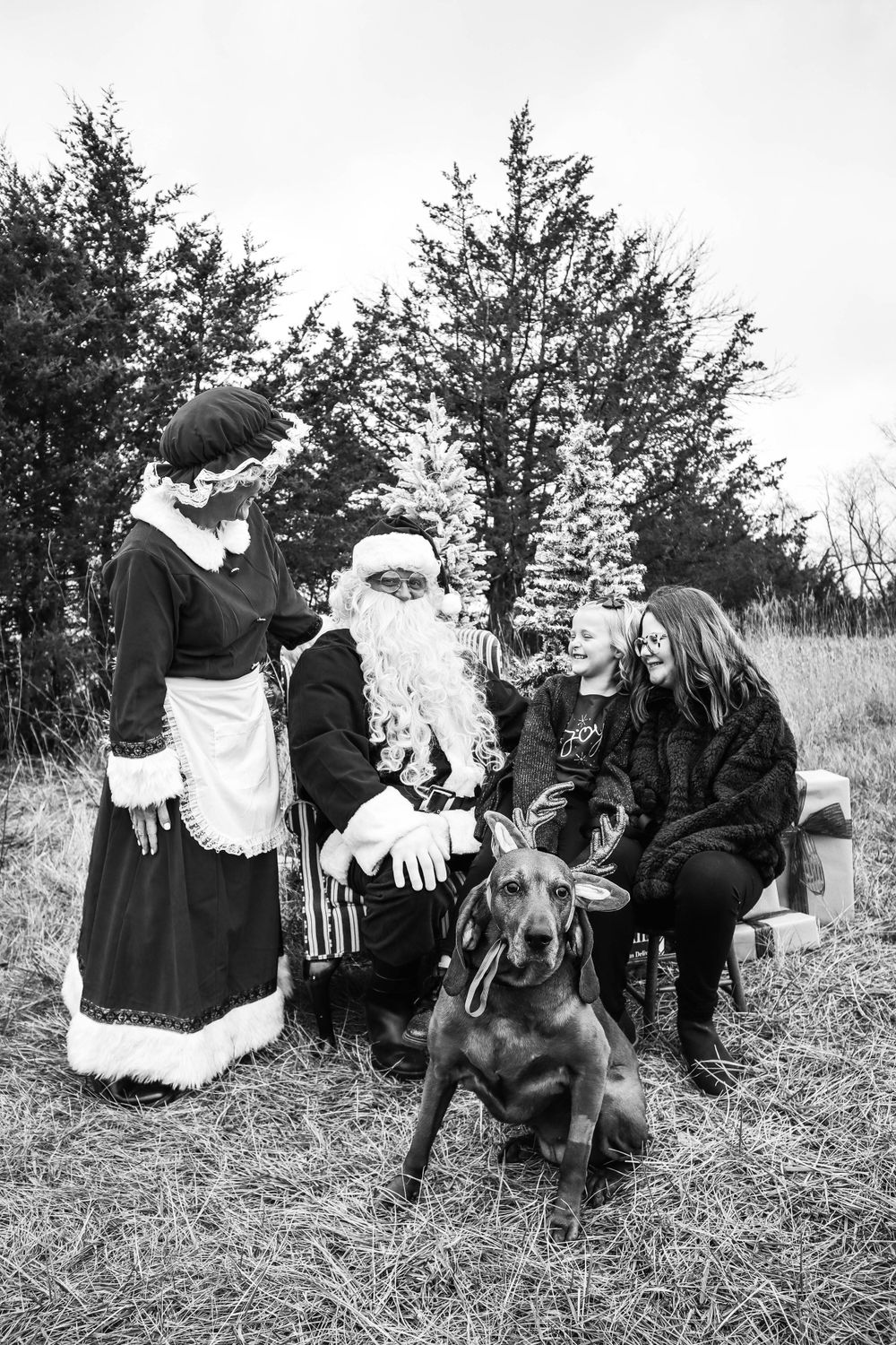 A black and white photo of Santa, Mrs. Claus, and others with a large dog in an outdoor setting.
