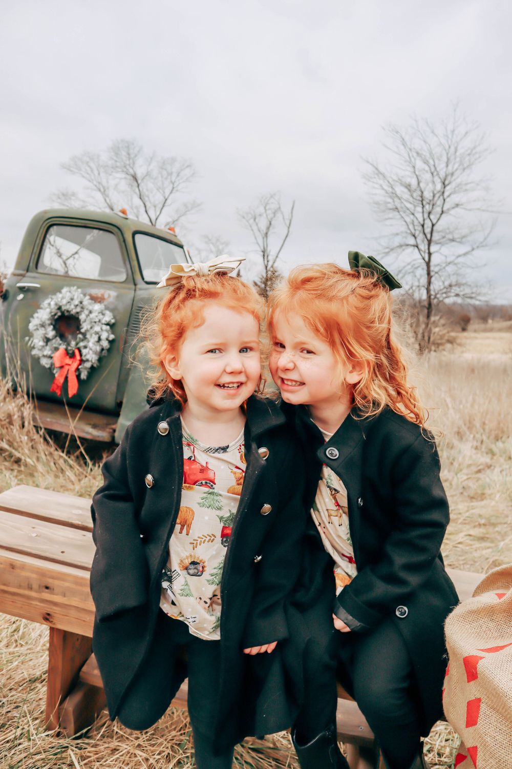 Two children in matching dark coats pose in front of a vintage truck decorated with a holiday wreath.