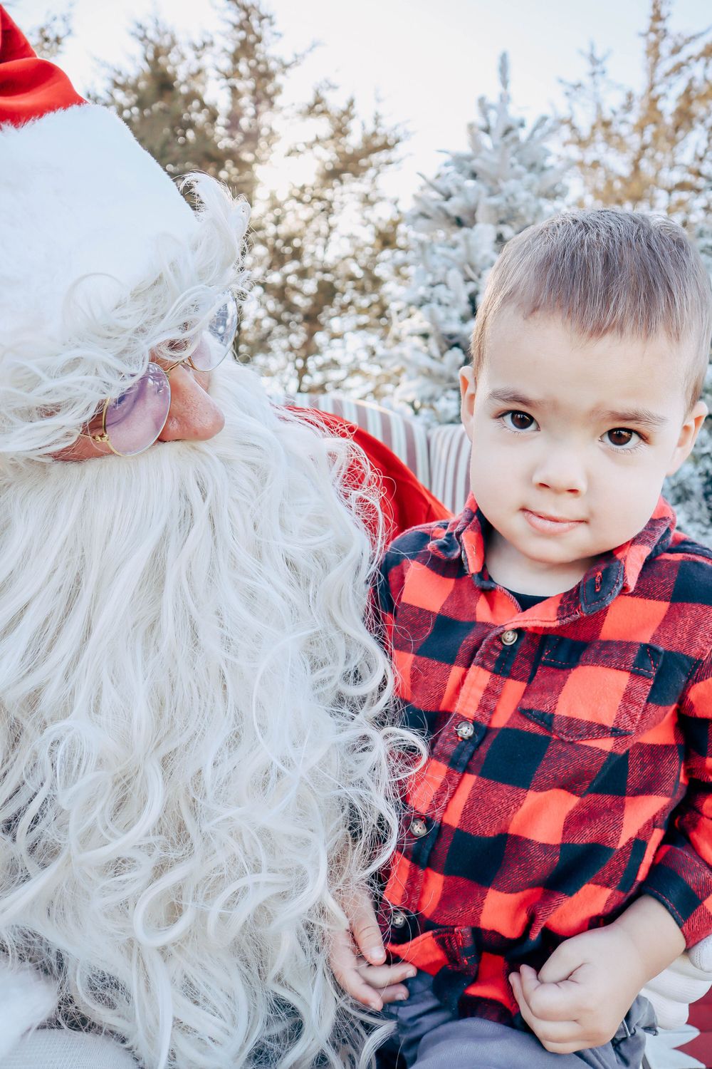 Santa Claus in traditional red suit poses with a small child wearing a buffalo plaid shirt in a winter setting.