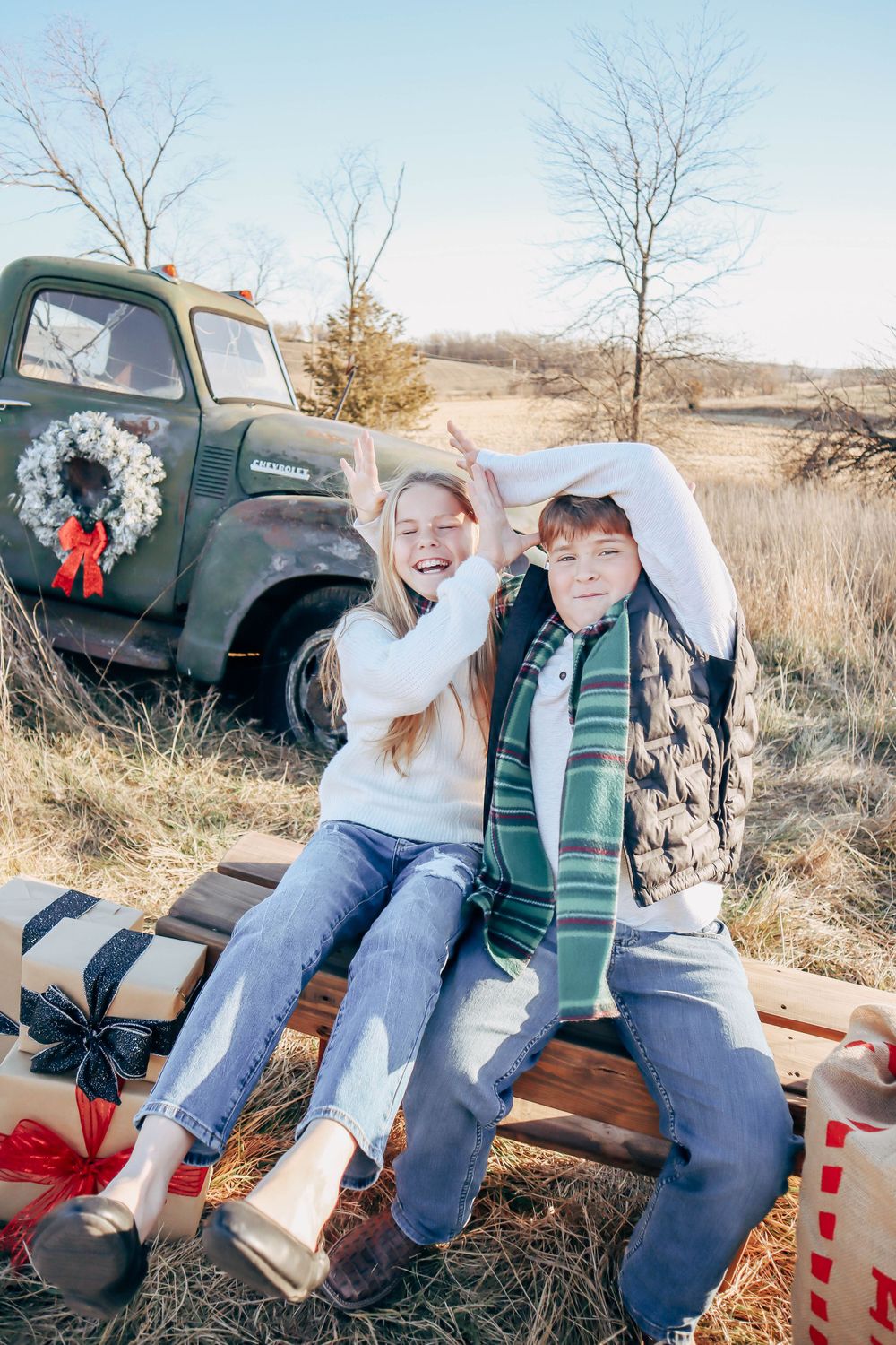 Two children sit on a wooden bench in front of a vintage green truck decorated with a Christmas wreath.