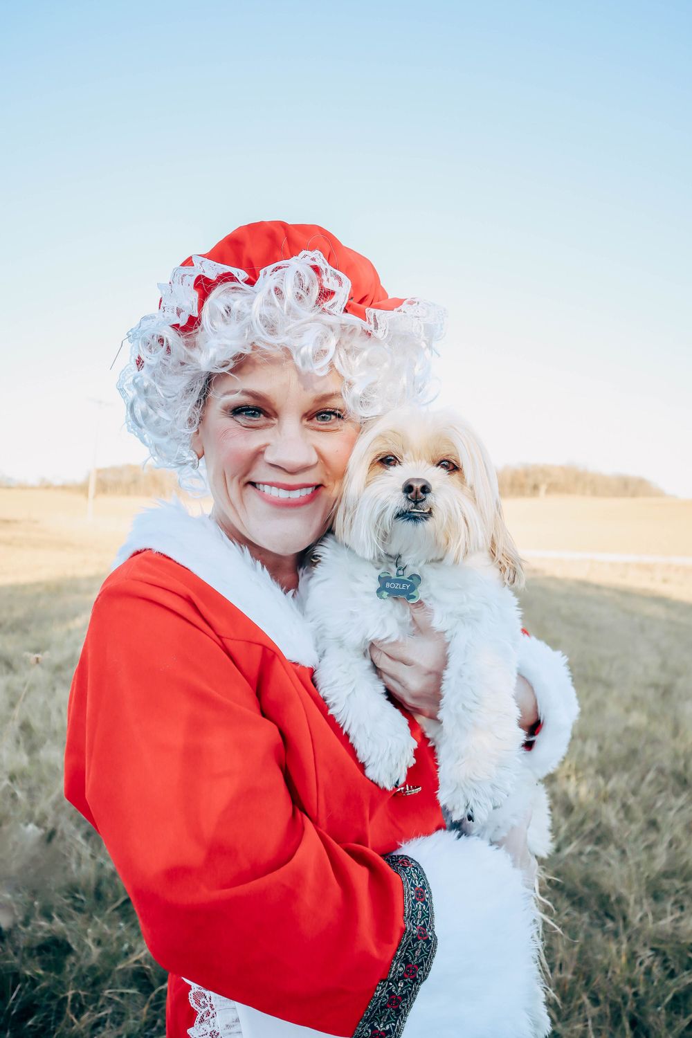 Mrs. Claus in red attire holding a small white fluffy dog outdoors on a sunny winter day.