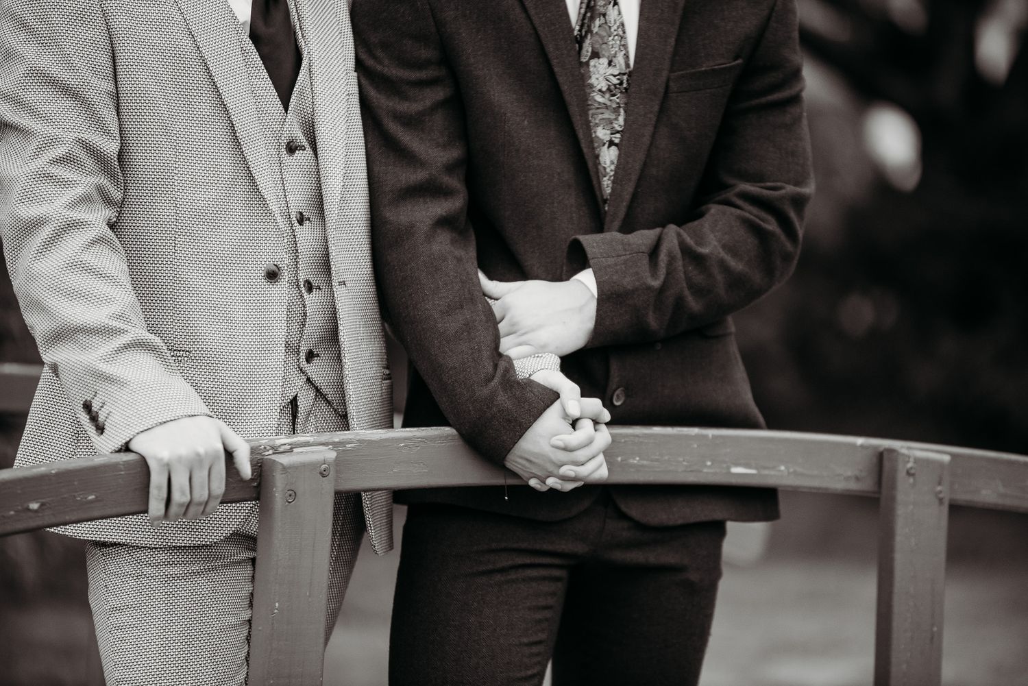 Close-up shot of two people holding hands on a wooden railing, wearing contrasting formal suits.