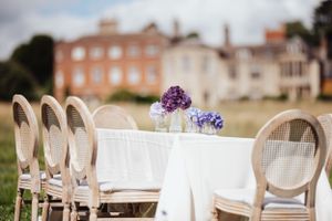 Elegant outdoor table setting with vintage chairs and purple floral centerpieces in front of a historic brick mansion.