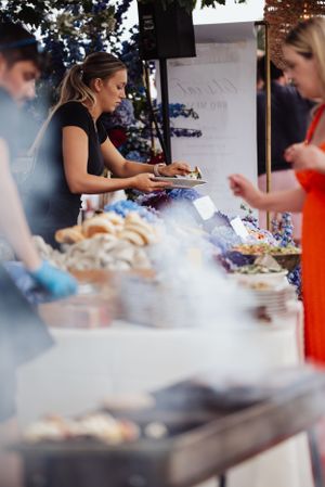 Food service staff preparing dishes at an outdoor catering buffet table with various menu items.