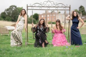 Four women in formal dresses pose together on a grassy field with a decorative metal arch frame.