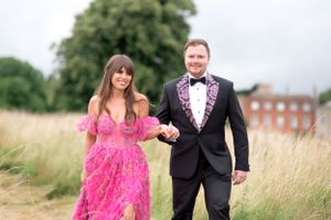 A couple in formal attire walking through a tall grass field with a brick building in the background.