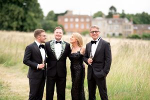 A group in formal black tie attire poses together in a grassy field with a brick mansion in the background.