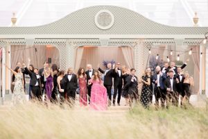 A large wedding party poses together in front of an elegant white gazebo with tall grass in the foreground.