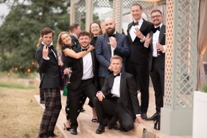 A wedding party in black tuxedos and formal attire poses playfully together outside a decorative gazebo structure.