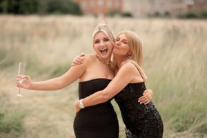 Two friends in black dresses share a joyful moment while taking photos in a tall grass field.