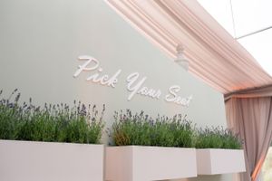 A neon sign reading 'Pick Your Seat' mounted above white planters filled with lavender flowers against a blush pink wall.