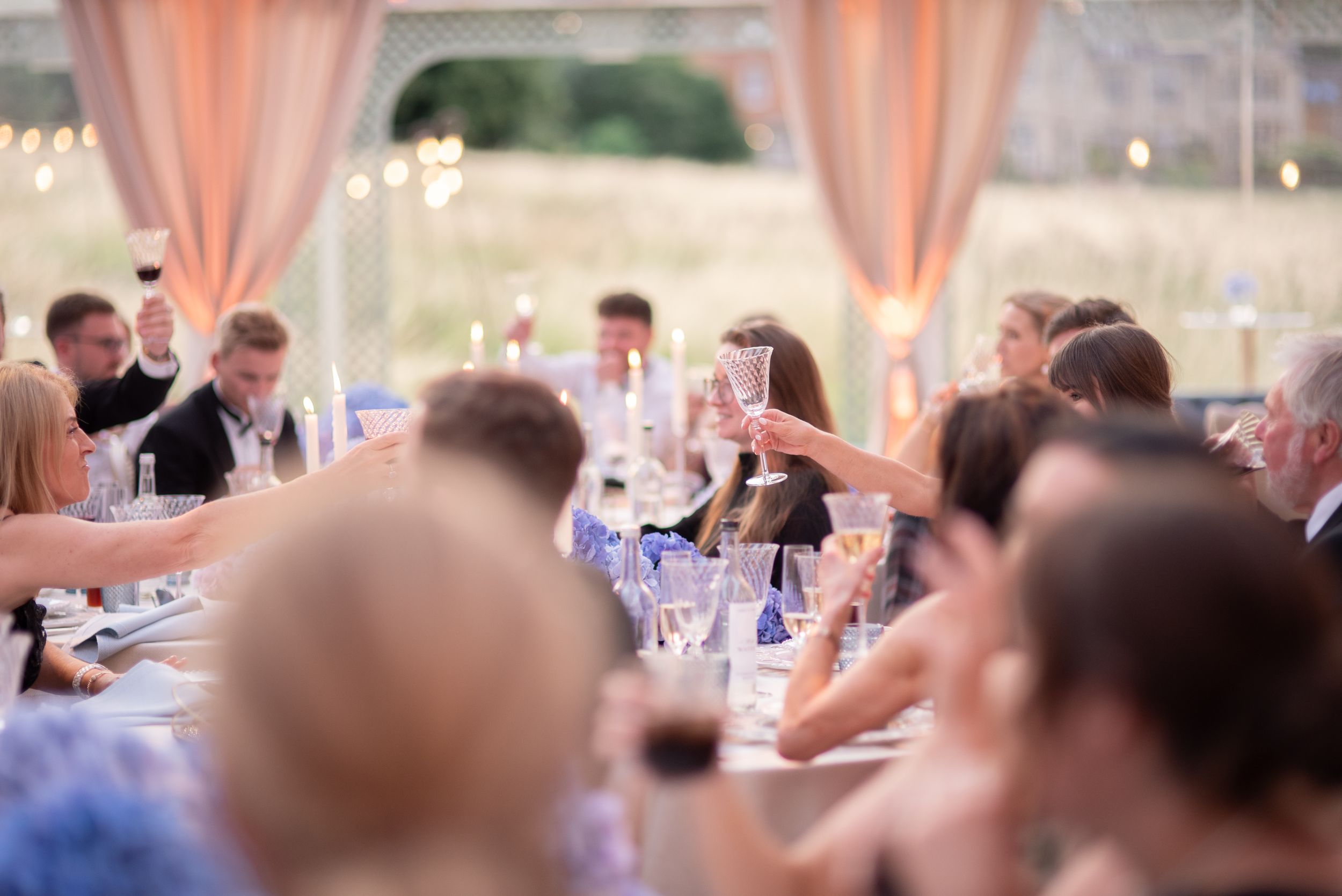 Wedding guests enjoy dinner and conversation at an elegant reception with soft pink drapes and ambient lighting.