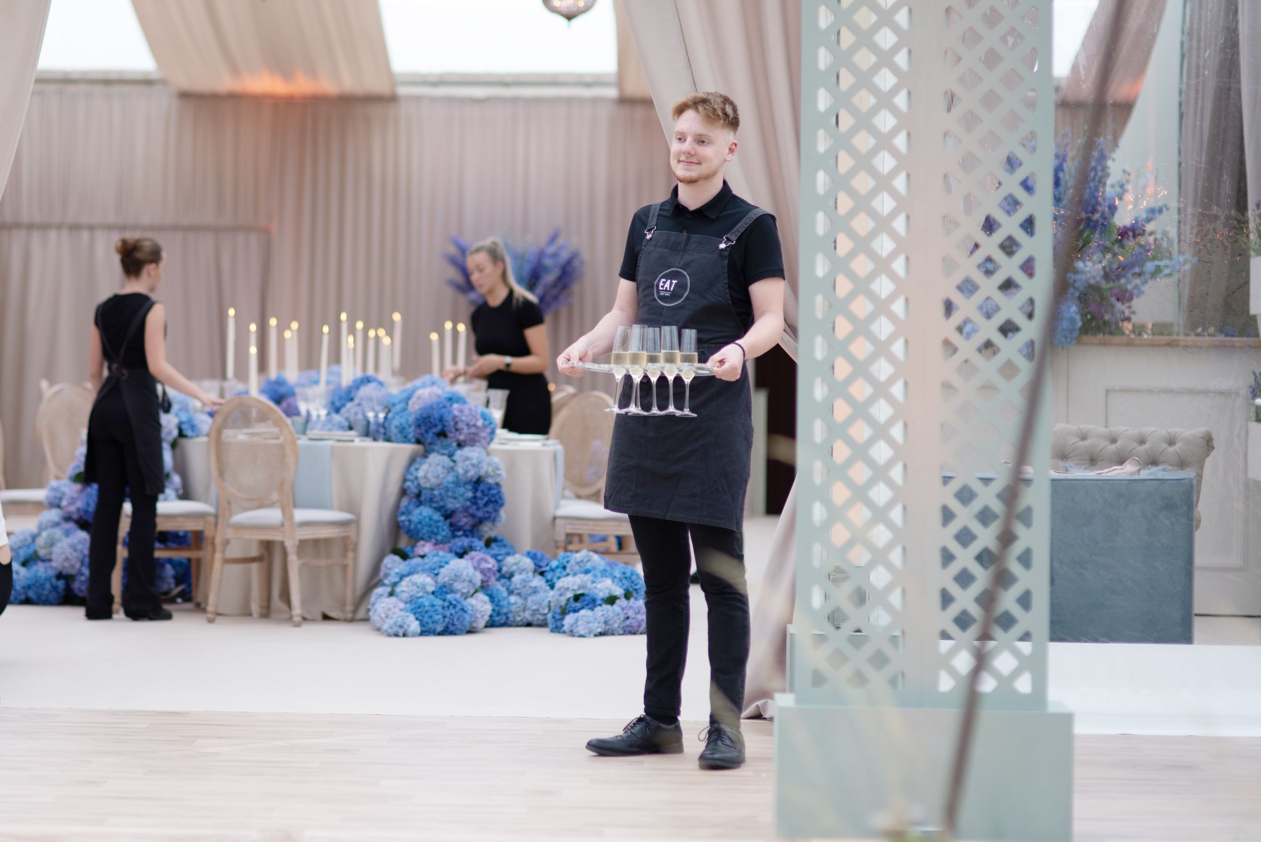 Service staff member in black uniform carrying champagne flutes at an elegant event venue with blue decor.