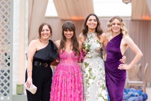 Four women pose together at a formal event wearing elegant dresses in black, pink, floral, and purple.
