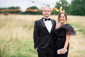 Elegant couple in formal attire standing in a field with string lights and tall grass at sunset.