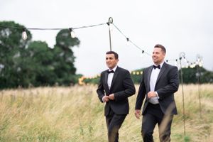 Two people in black tuxedos walking through a grassy field with string lights hanging overhead.