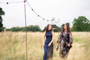 Two people stand together in a tall grass field under string lights on a summer evening.
