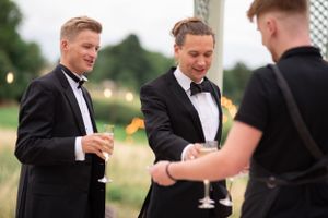Two people in black tuxedos and bow ties being served drinks by a server at an outdoor event.