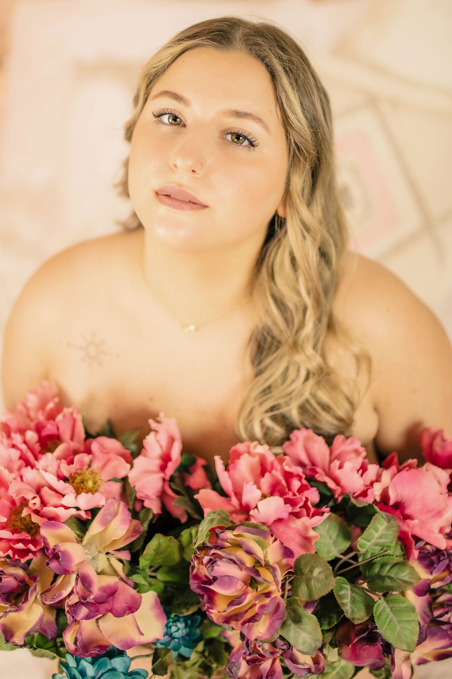 Curvy blonde woman in light pink lingerie holding flowers over her chest in a soft pastel studio boudoir session, embracing self-love.