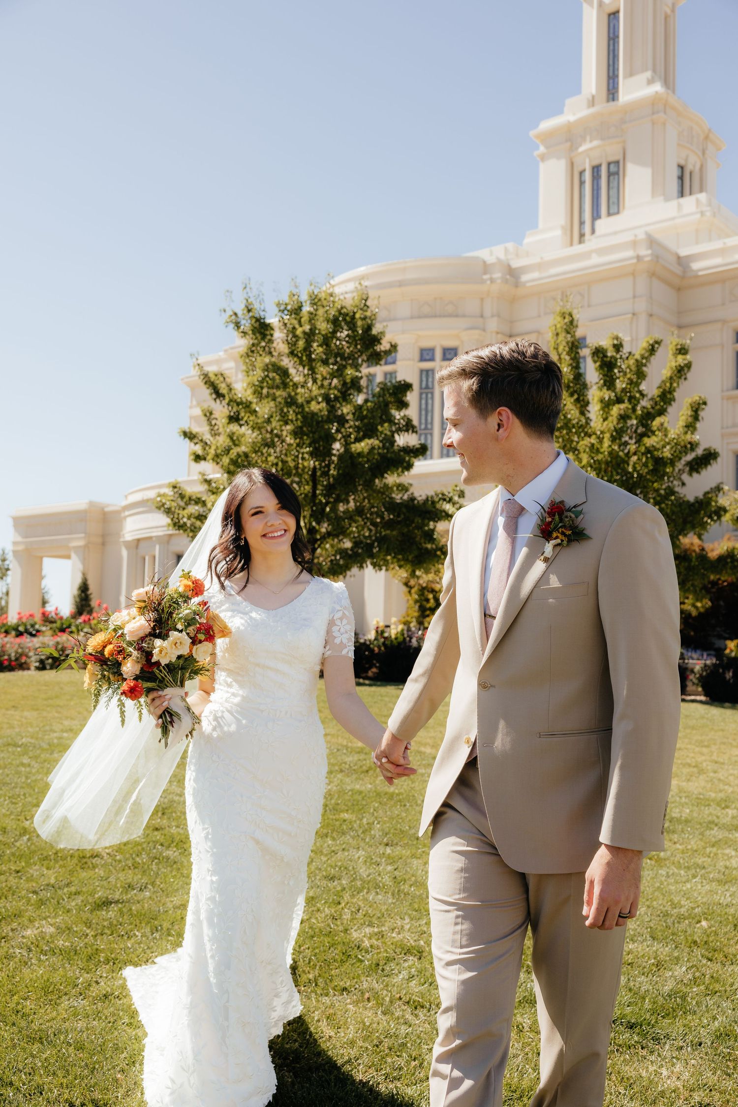 Bride and groom holding hands and smiling outside a white LDS temple on a sunny day.