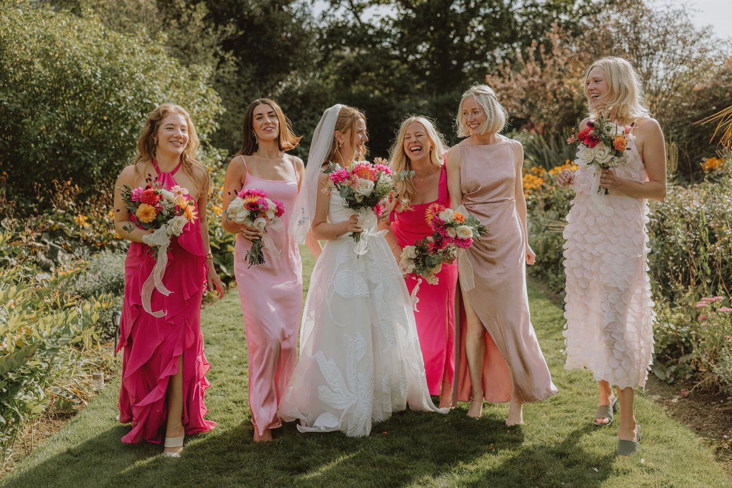 Bridal party wearing various shades of pink dresses poses together in garden setting.