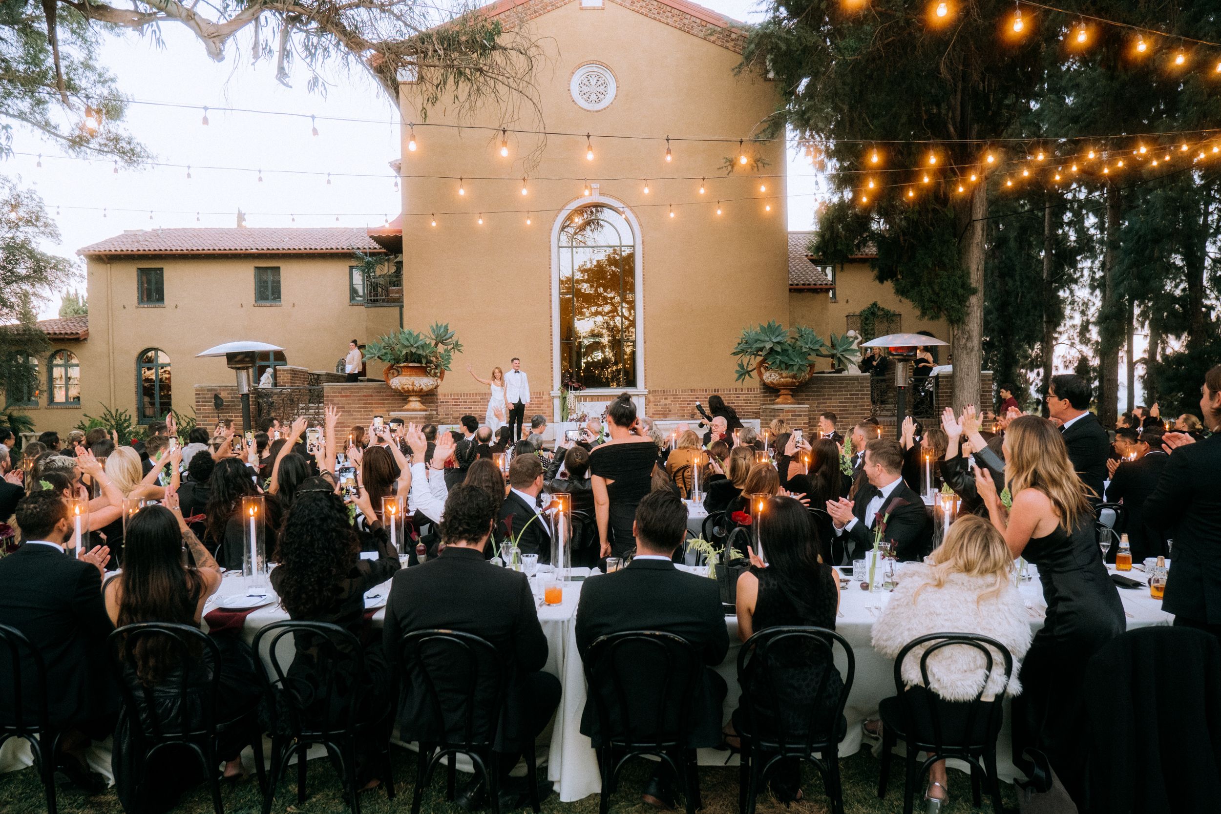 Large group dining at outdoor wedding reception with string lights and Mediterranean-style building.