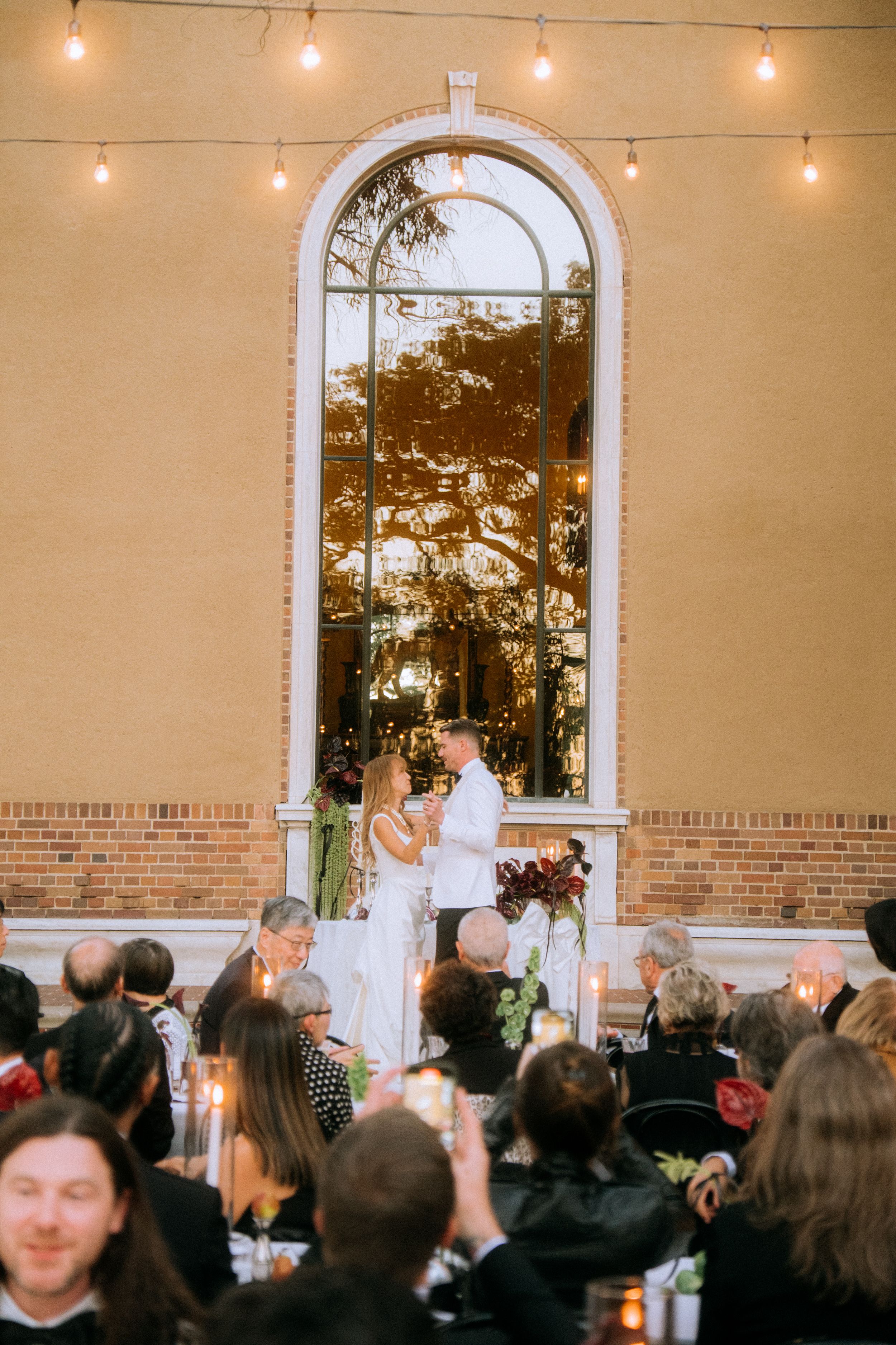 Wedding celebration under arched window with string lights and gathered guests at outdoor reception.