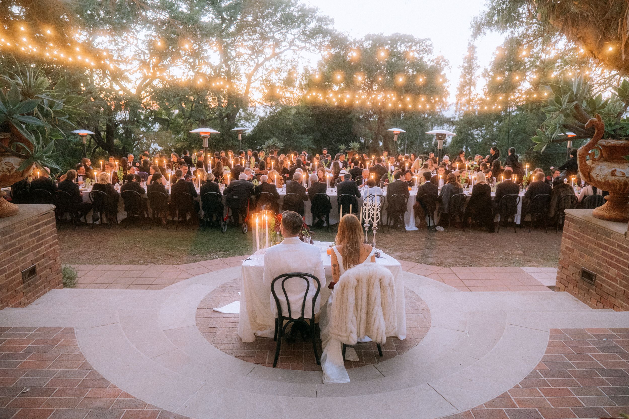 Outdoor wedding reception at dusk with string lights and large crowd facing a couple.