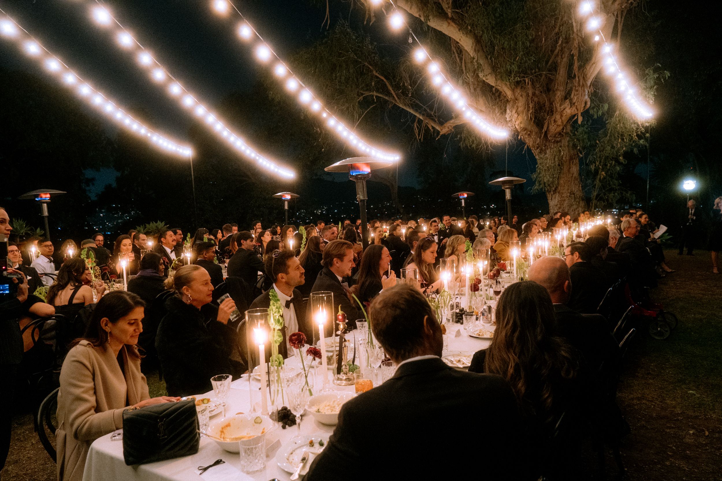 Long dinner table under string lights at night with guests enjoying an outdoor celebration.