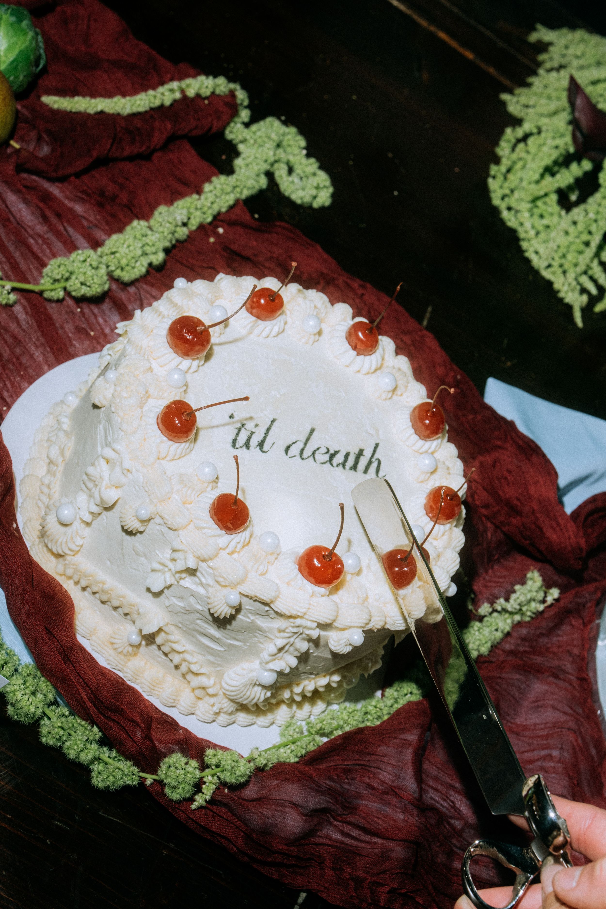 White frosted cake decorated with cherries and 'til death' text on burgundy fabric.