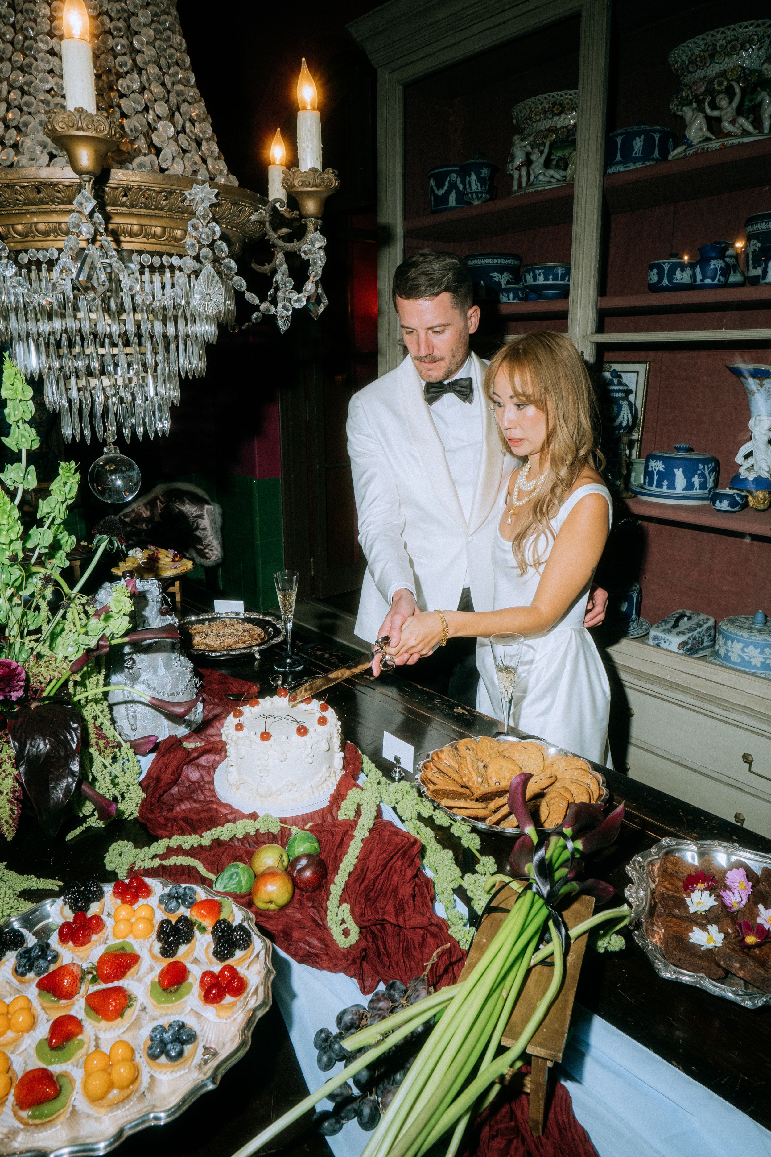Wedding couple cutting cake at elegant dessert table with crystal chandelier and appetizers.