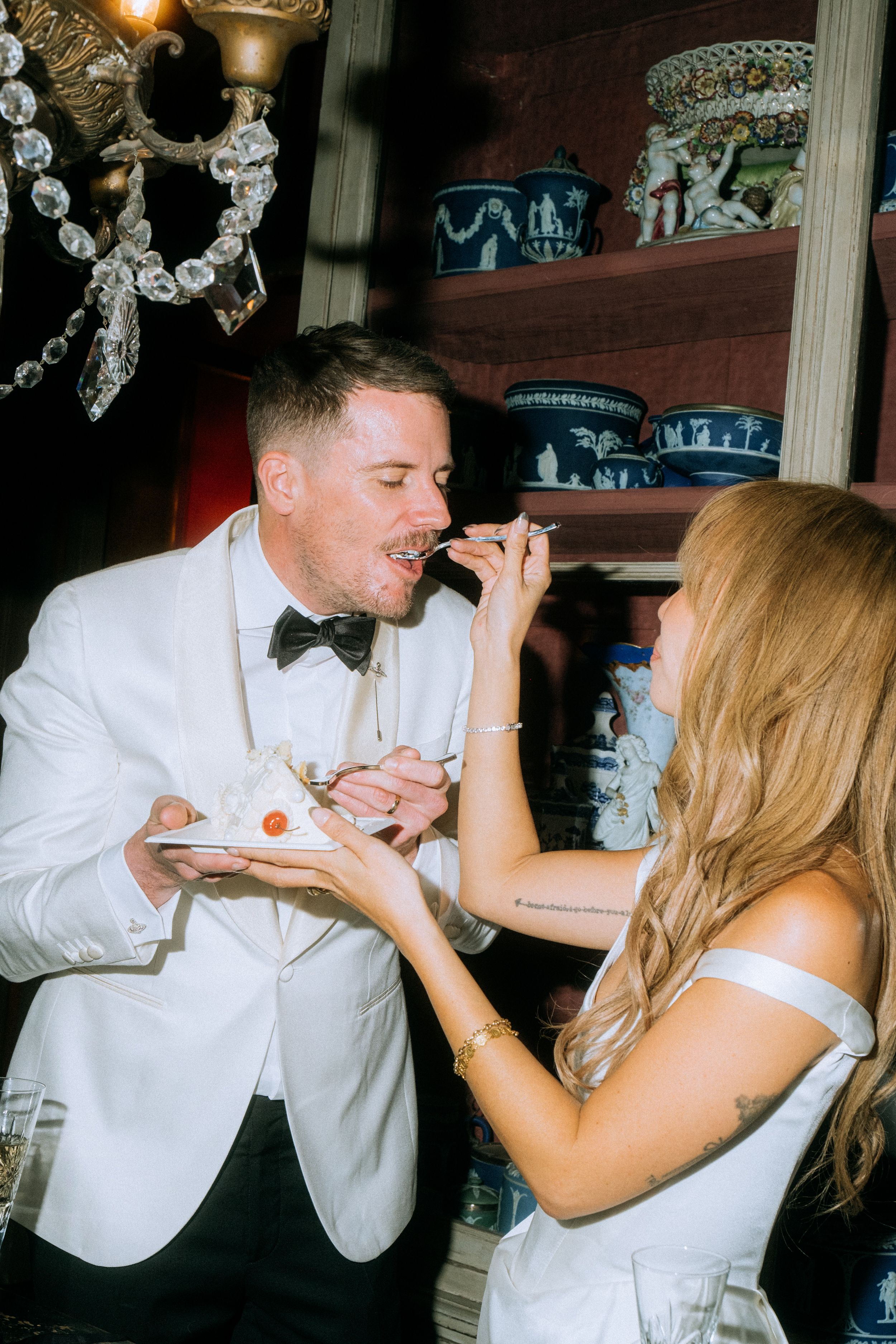 Couple sharing wedding cake in formal attire under crystal chandelier.