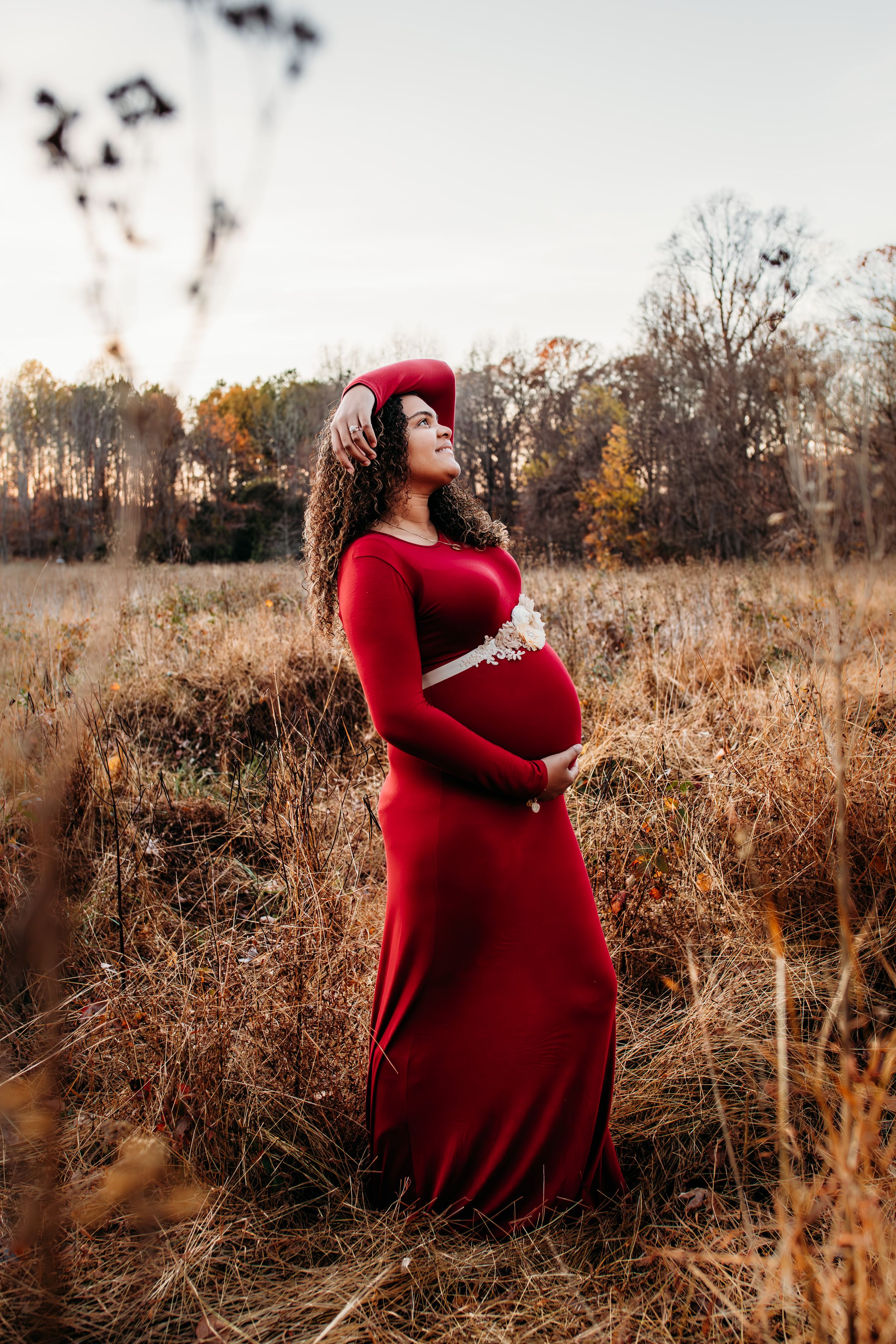 Maternity woman standing in field in red dress for maternity photography. Captured by Maryland Photographer Mallory Bramlett.