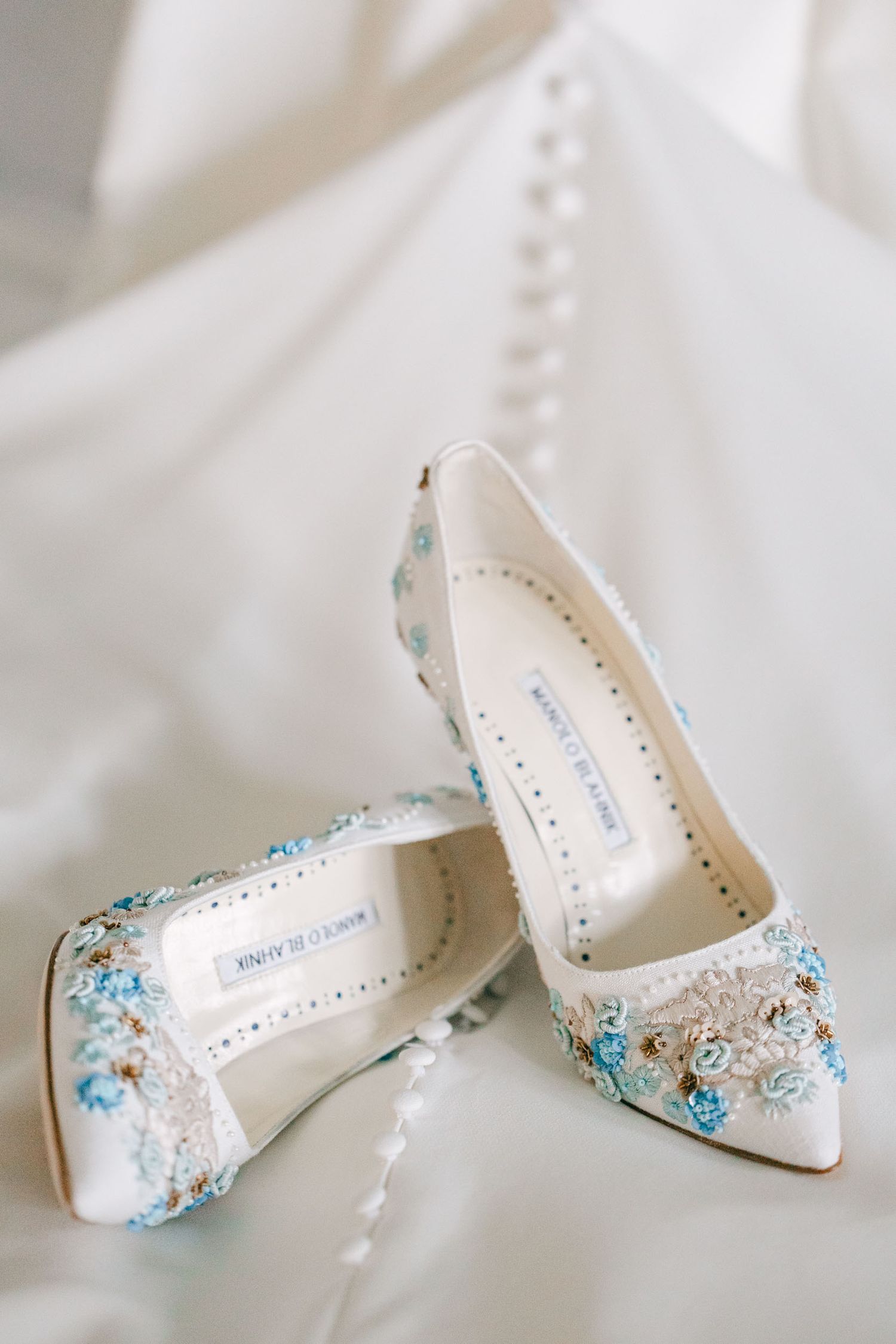 Elegant white bridal shoes with delicate blue floral embellishments and crystal details on a light fabric background.