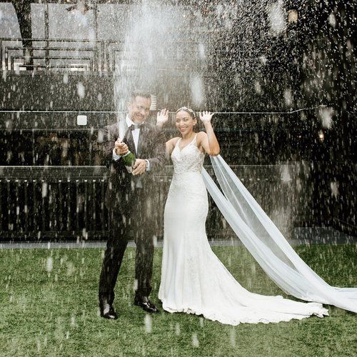 A couple in wedding attire poses together in the rain, with water droplets falling around them in a garden setting.