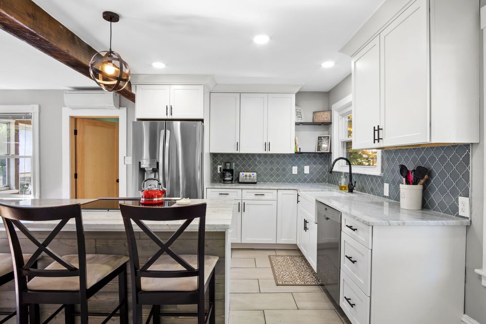 Modern white kitchen with gray backsplash, cross-back chairs, and stainless steel appliances.