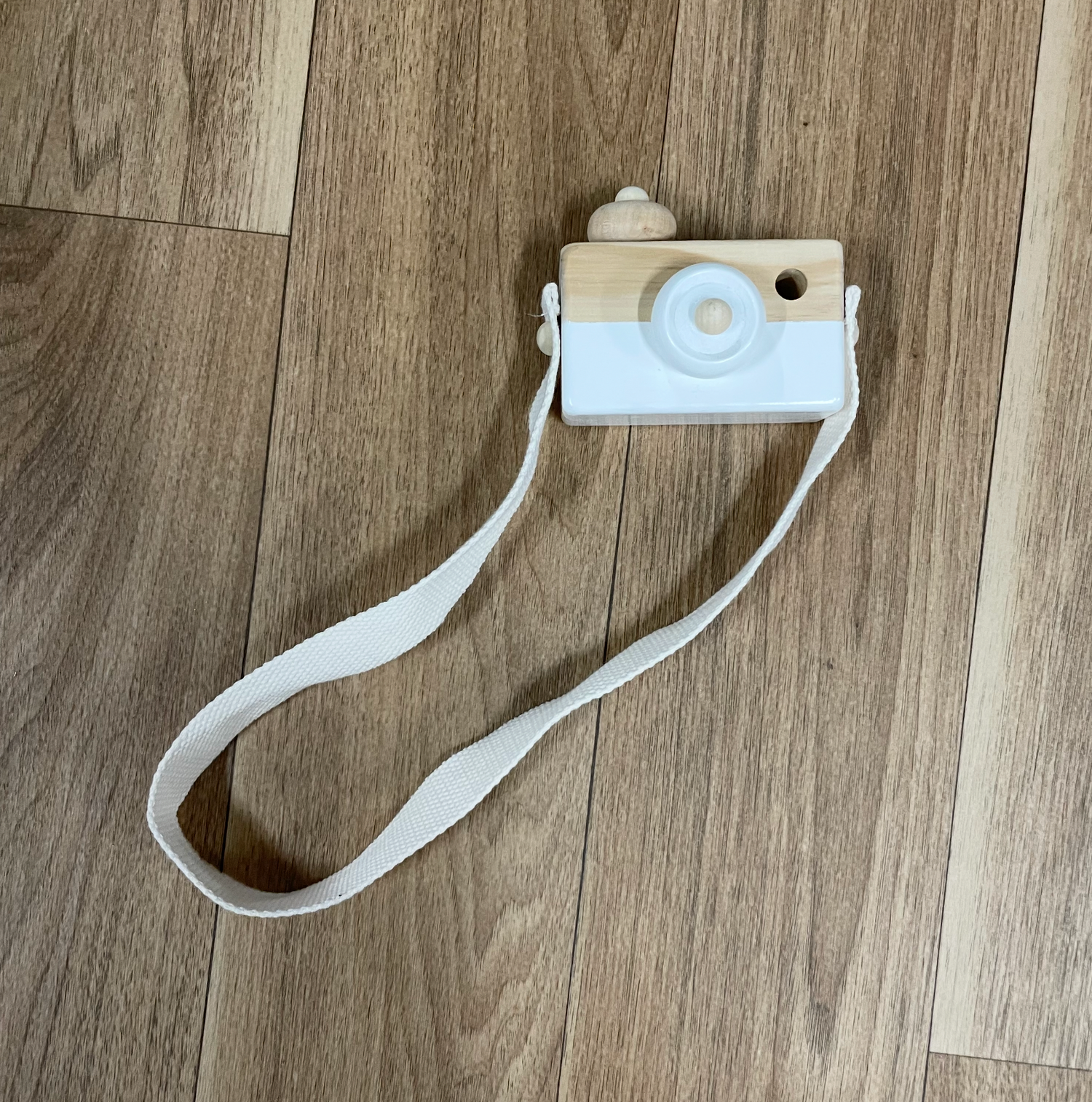 A white wooden toy camera with strap lying on hardwood flooring.