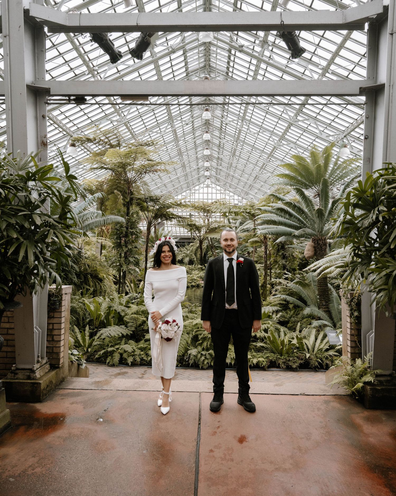 A couple standing in a bright conservatory with glass ceilings and tropical plants, creating an elegant greenhouse setting.