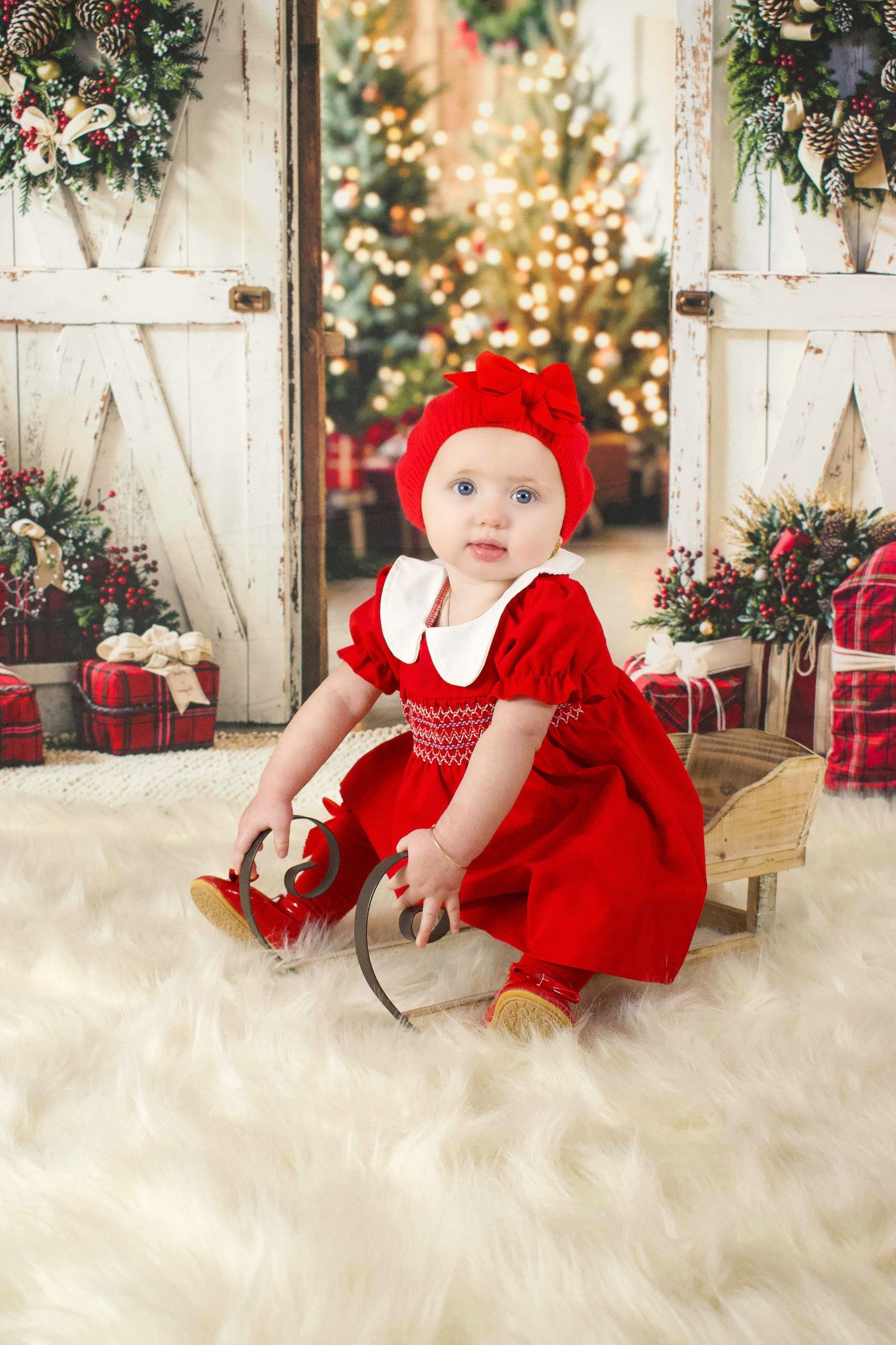 Baby in red dress sitting on white fur rug with Christmas backdrop and decorations.