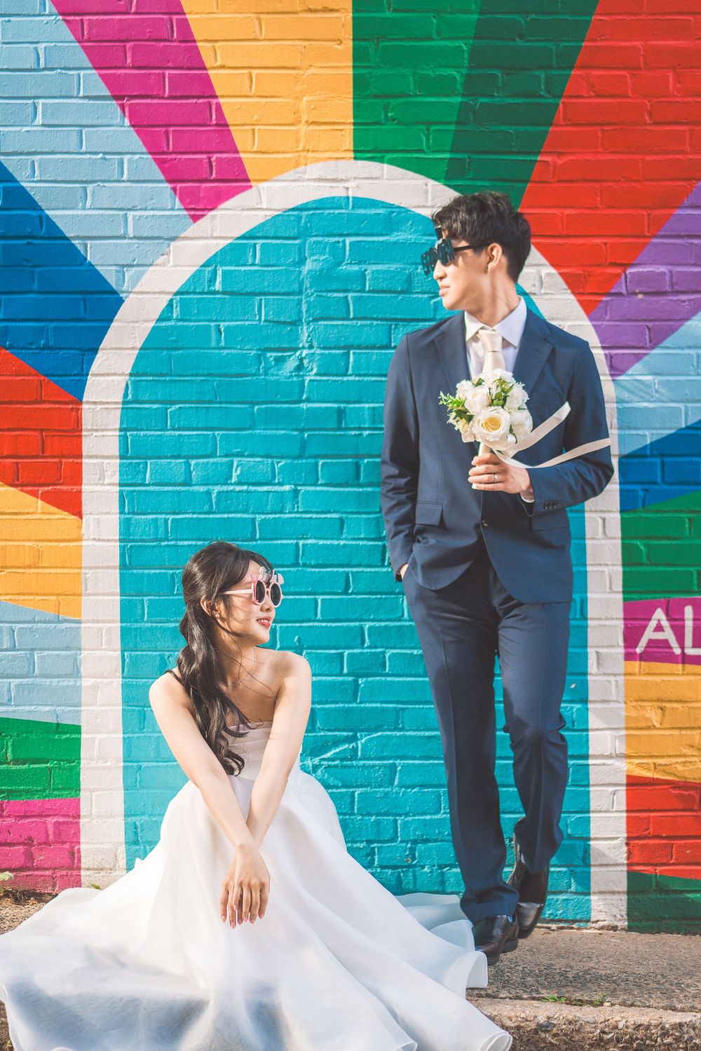 Wedding couple poses in front of a colorful geometric rainbow mural painted on brick wall.