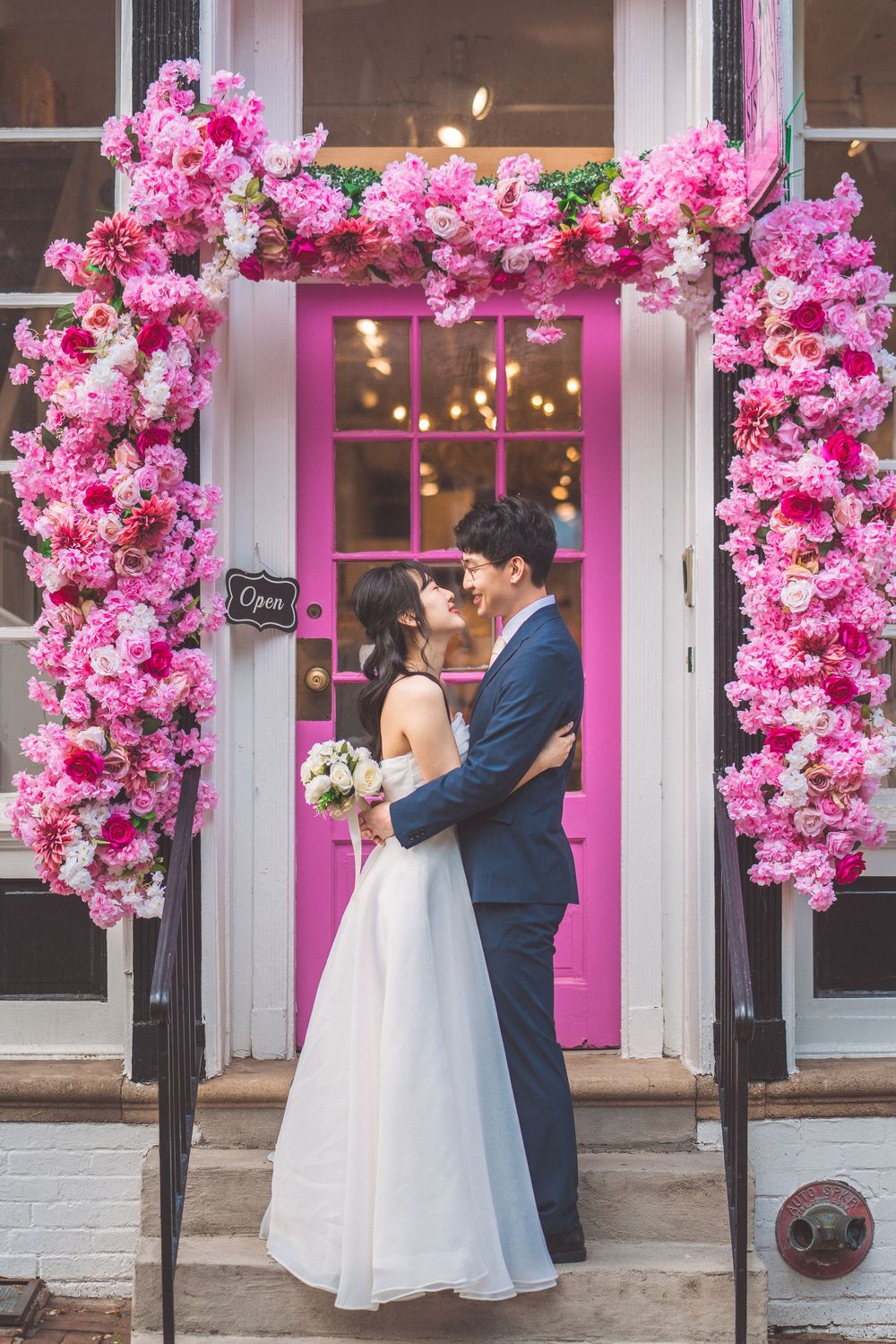 A romantic wedding couple stands under a pink floral archway at a storefront entry with a bright pink door.
