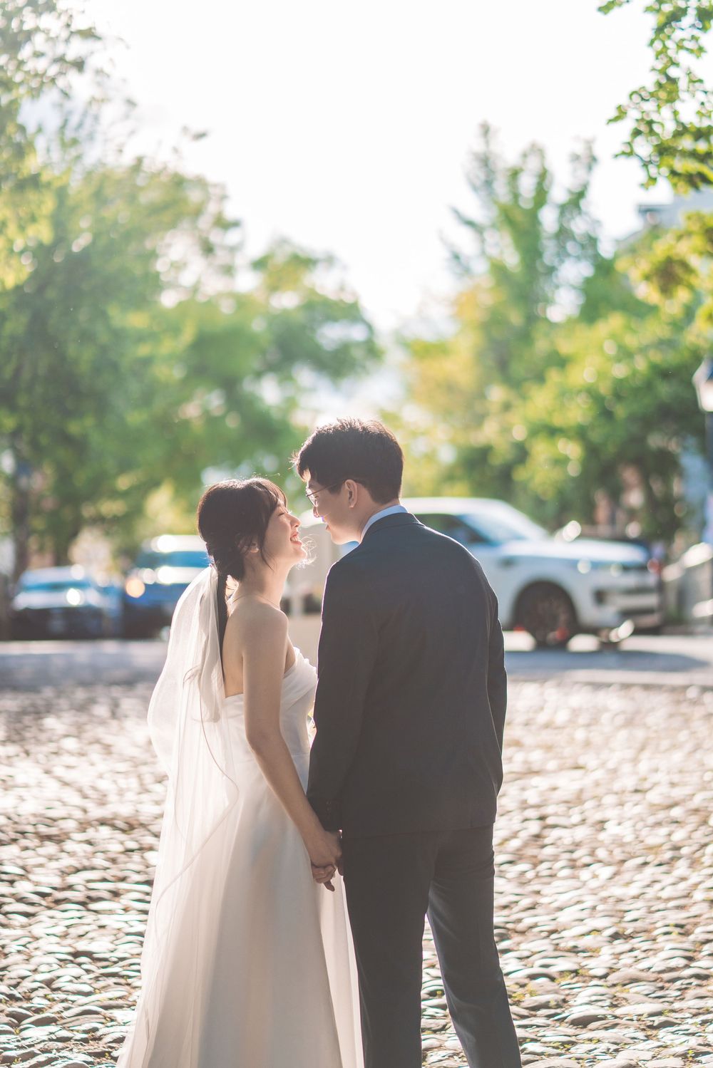 A romantic wedding couple shares an intimate moment together on a cobblestone street as sunlight filters through trees.