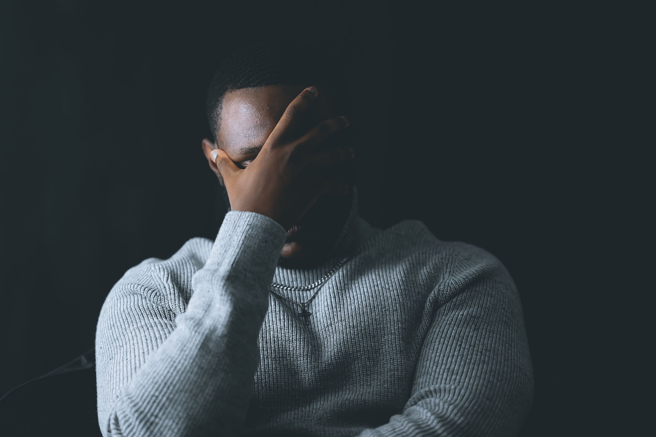 Person in light gray sweater covering face with hand against dark background in moody portrait.