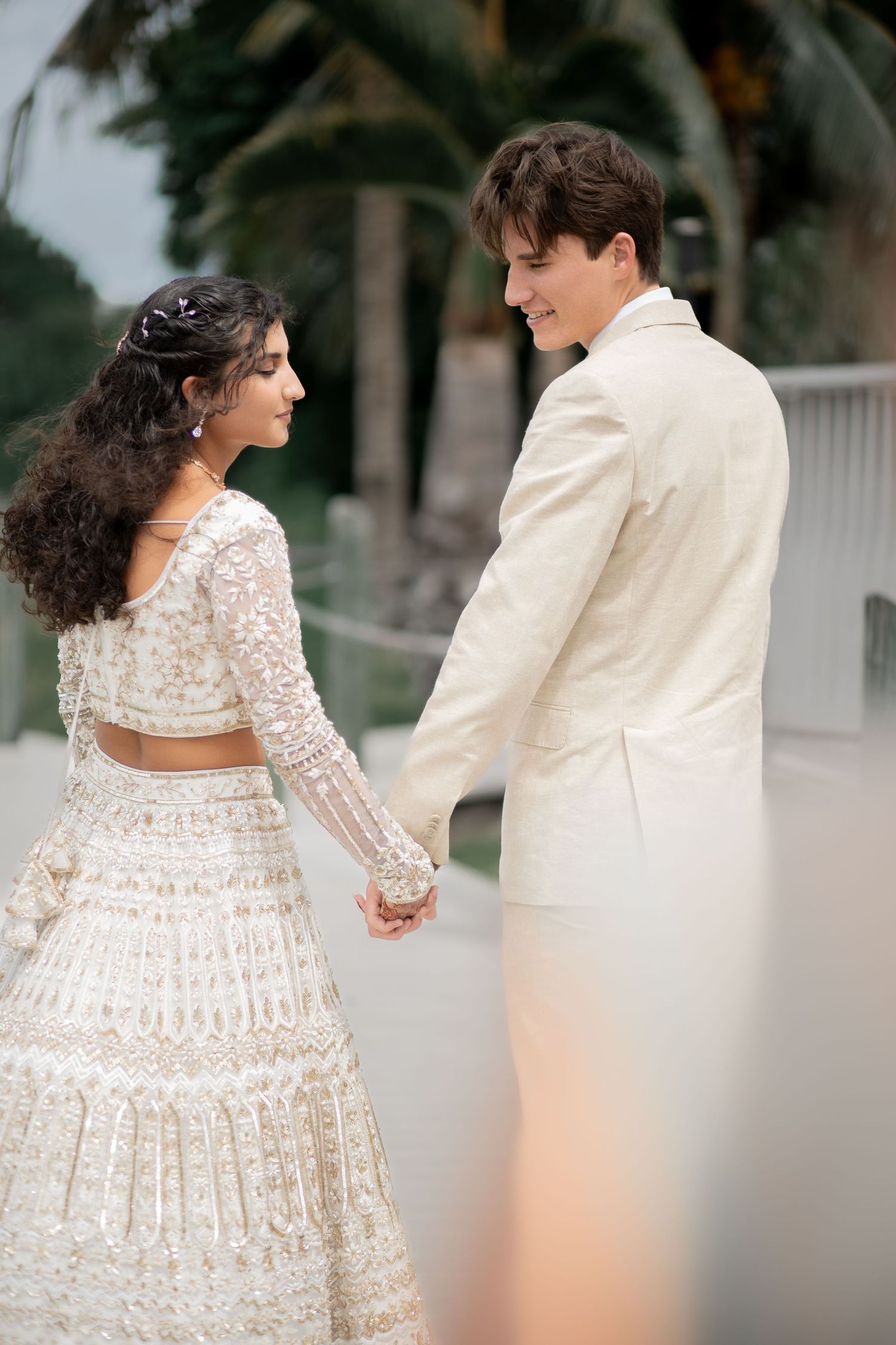 Couple in elegant white wedding attire holding hands outdoors with palm trees in soft romantic lighting.