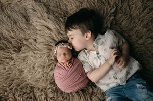 A newborn baby wrapped in pink fabric lies on a fuzzy beige rug next to an older sibling.