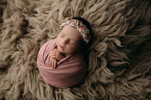 Sleeping newborn wrapped in mauve swaddle wearing floral headband on fluffy grey textured blanket.